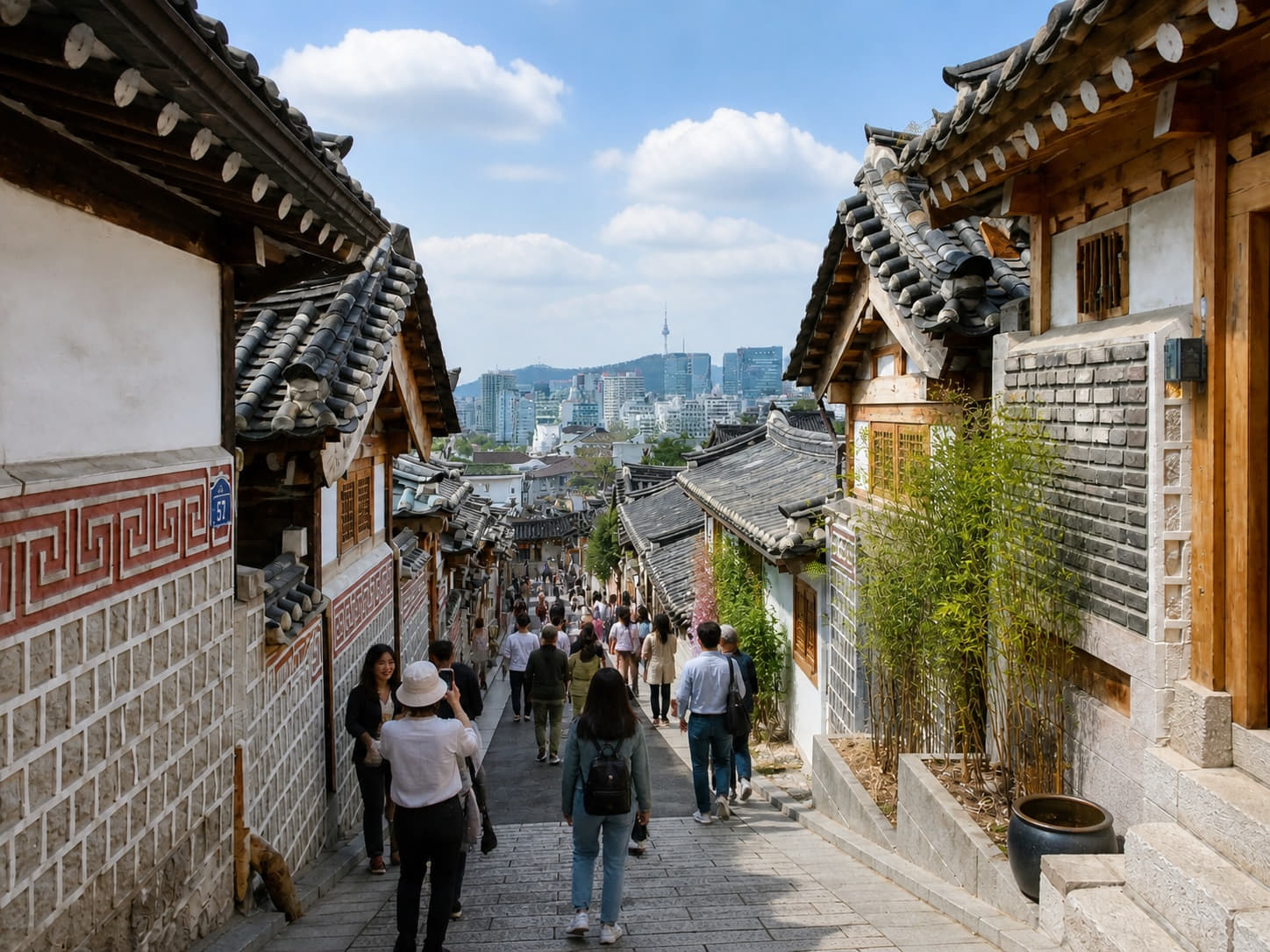 A scenic view of a narrow alley in Bukchon Hanok Village, Seoul, featuring traditional Korean houses with tiled roofs and the N Seoul Tower in the distant background under a blue sky